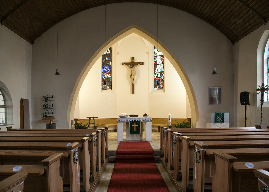 Hinter dem Spitzbogen ist der Chorraum mit Holzbänken und Kreuz sowie Buntglasfenstern